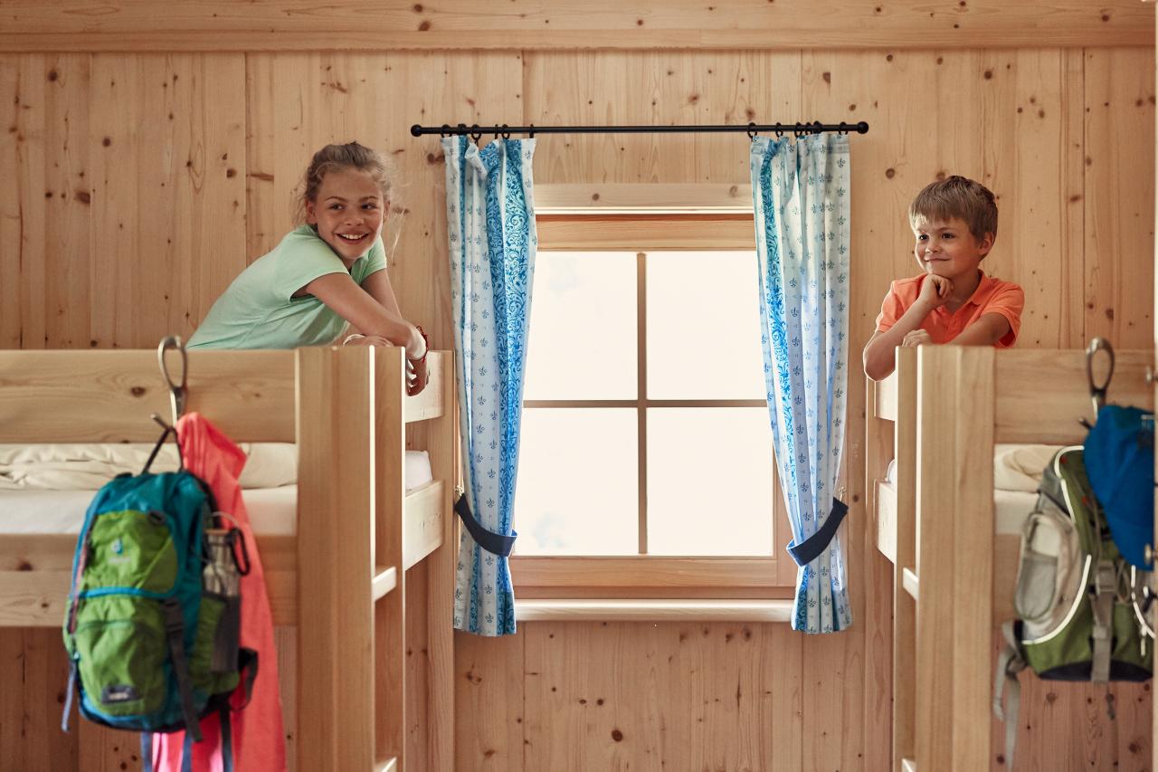 Dvě děti sedí na palandách v pokoji obloženém dřevem.