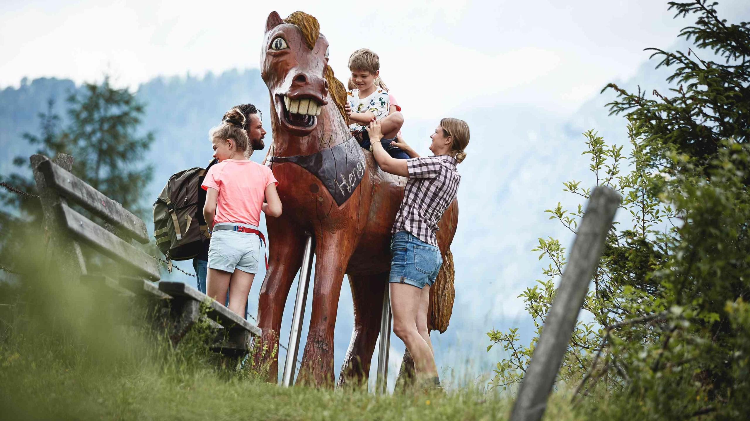 Dítě sedí na dřevěném koni v životní velikosti obklopené rodinou.