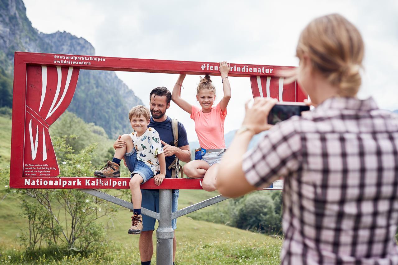 Žena fotící muže a dvě děti v selfie rámečku na alpské louce