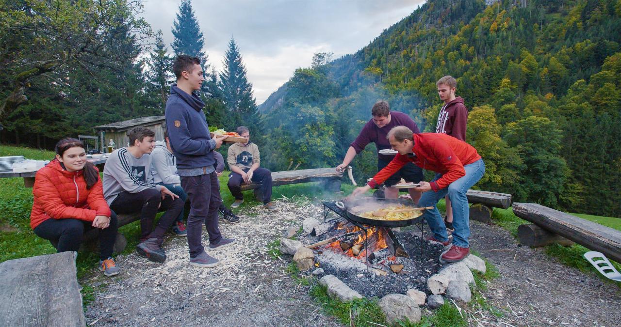 Mladí lidé sedí u ohně, na kterém se připravuje jídlo na pánvi.