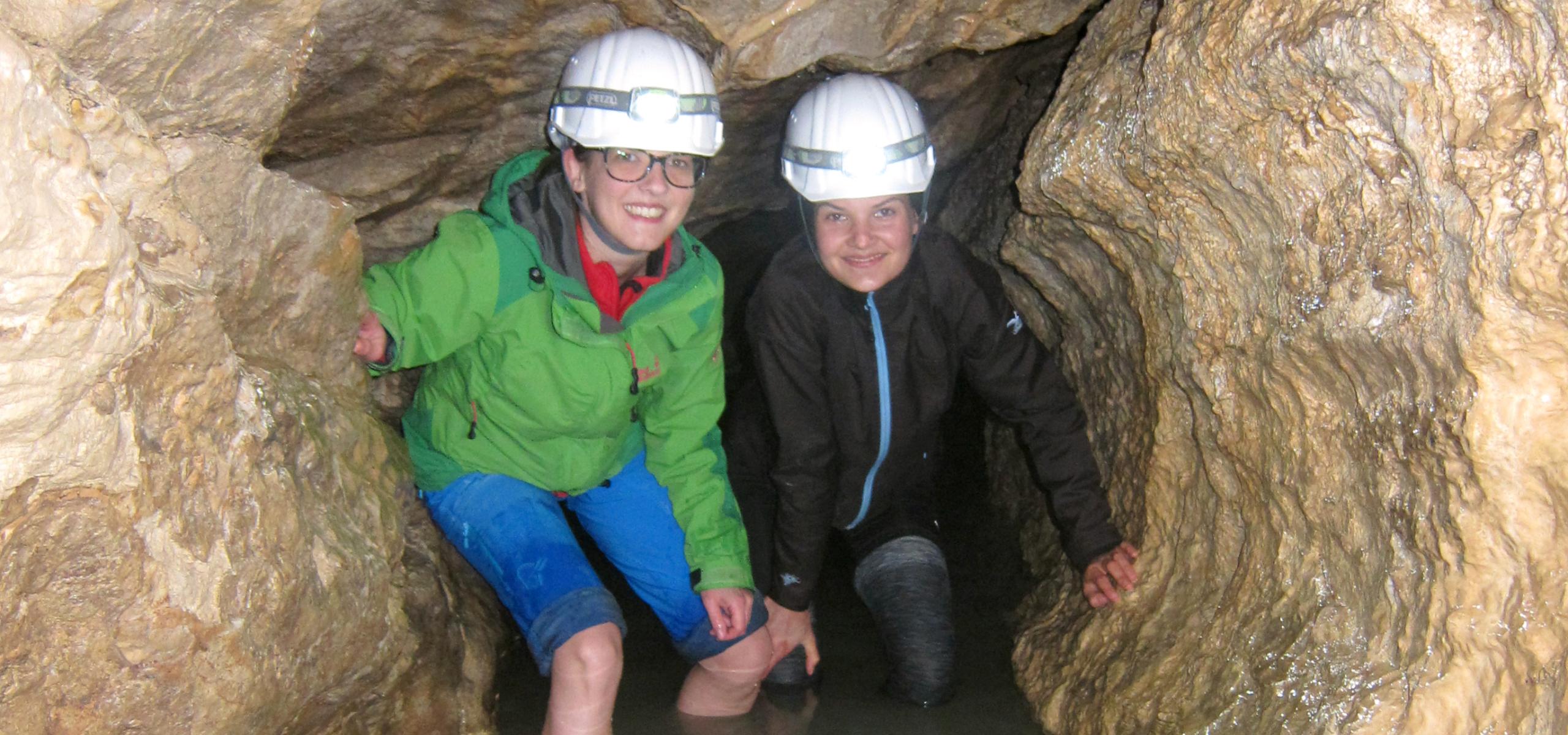 Dvě děti se brodí po kolena ve vodě v jeskynní chodbě.