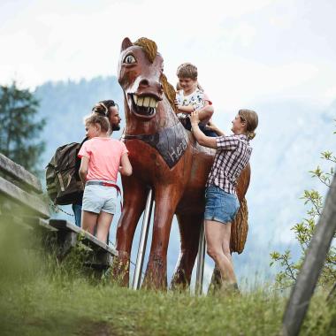 Dítě sedí na dřevěném koni v životní velikosti obklopené rodinou.