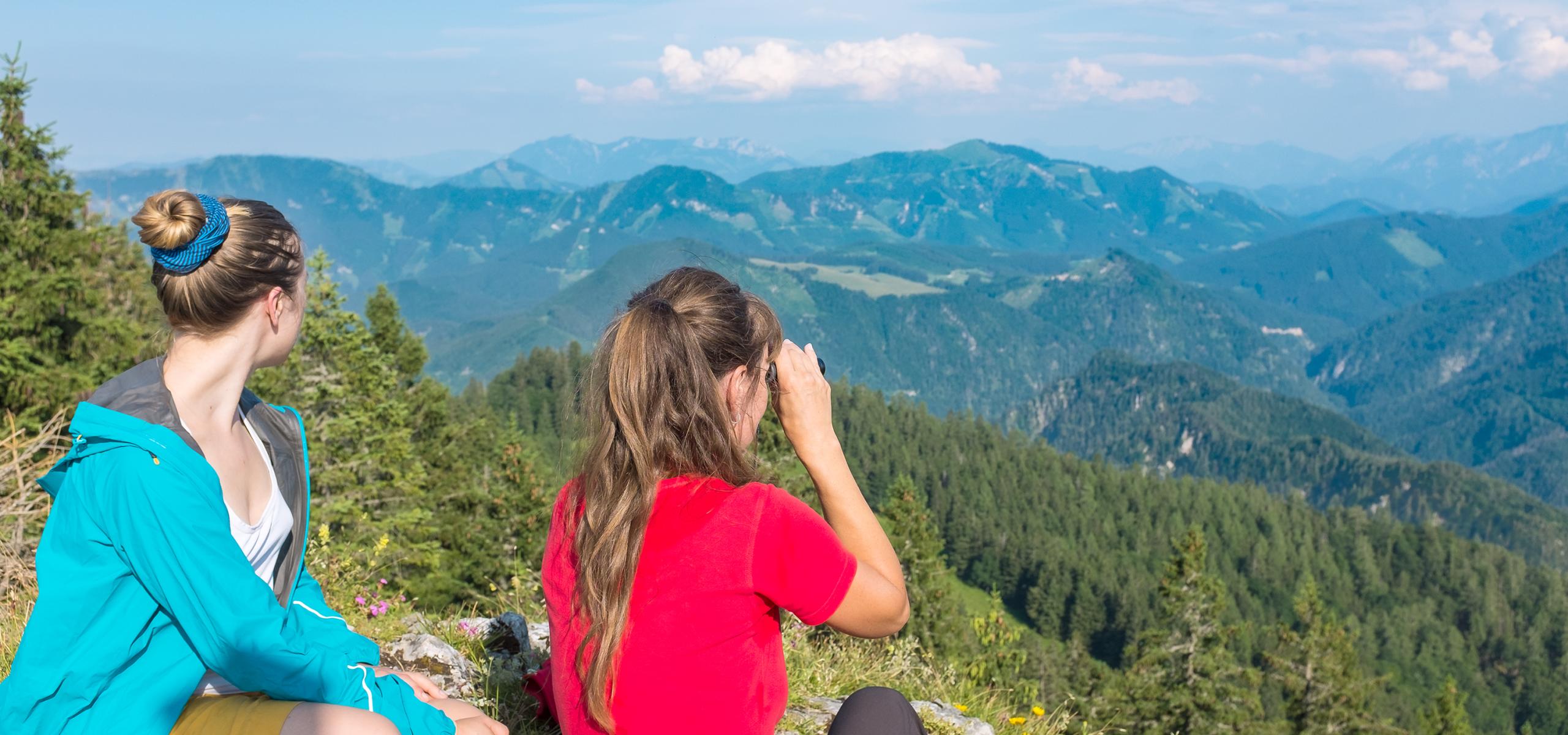 Dvě ženy sedí na vrcholu hory a pozorují horské panorama.
