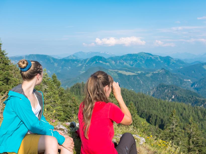 Dvě ženy sedí na vrcholu hory a pozorují horské panorama.